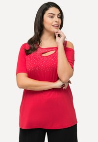 Rød kortærmet top med cold-shoulder design, der har en scoop neck og er udsmykket med metalliske nitter langs udskæringen.