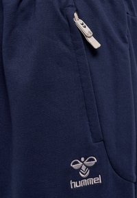 Marineblauwe sportbroek met een zachte textuur, voorzien van een zijzak en een ritsluiting. Hummel-logo geborduurd in lichtgrijs.