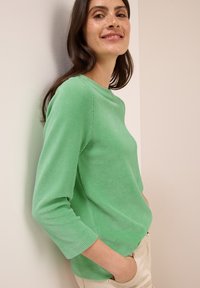 Lächelnde Frau mit dunklen Haaren, die ein grünes, geripptes Langarmoberteil und beige Hosen trägt, seitlich stehend mit Händen in den Taschen.