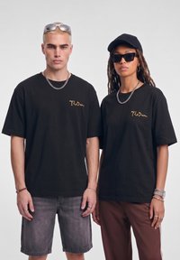 Δύο άτομα που φορούν μαύρα t-shirts με χρυσό κείμενο "The Dream", συνοδευμένα από αλυσίδες, δαχτυλίδια, γυαλιά ηλίου και casual σορτς και παντελόνια.