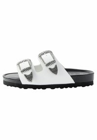 Vit halksandals med två justerbara remmar med intrikata silverfästen, en texturerad svart sula och ett slätt ovandelmaterial.