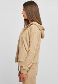 Beige zip-up hette med kengurulomme, snøring i hetten og ribbede ermer. Mykt stoff med en glatt tekstur. Modell vist i profil.
