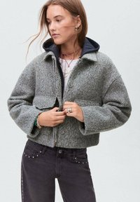 Veste en laine gris coupée avec fermeture à bouton, dotée de deux poches avant et d'une capuche bleu marine. Associée avec un jean noir orné de clous.