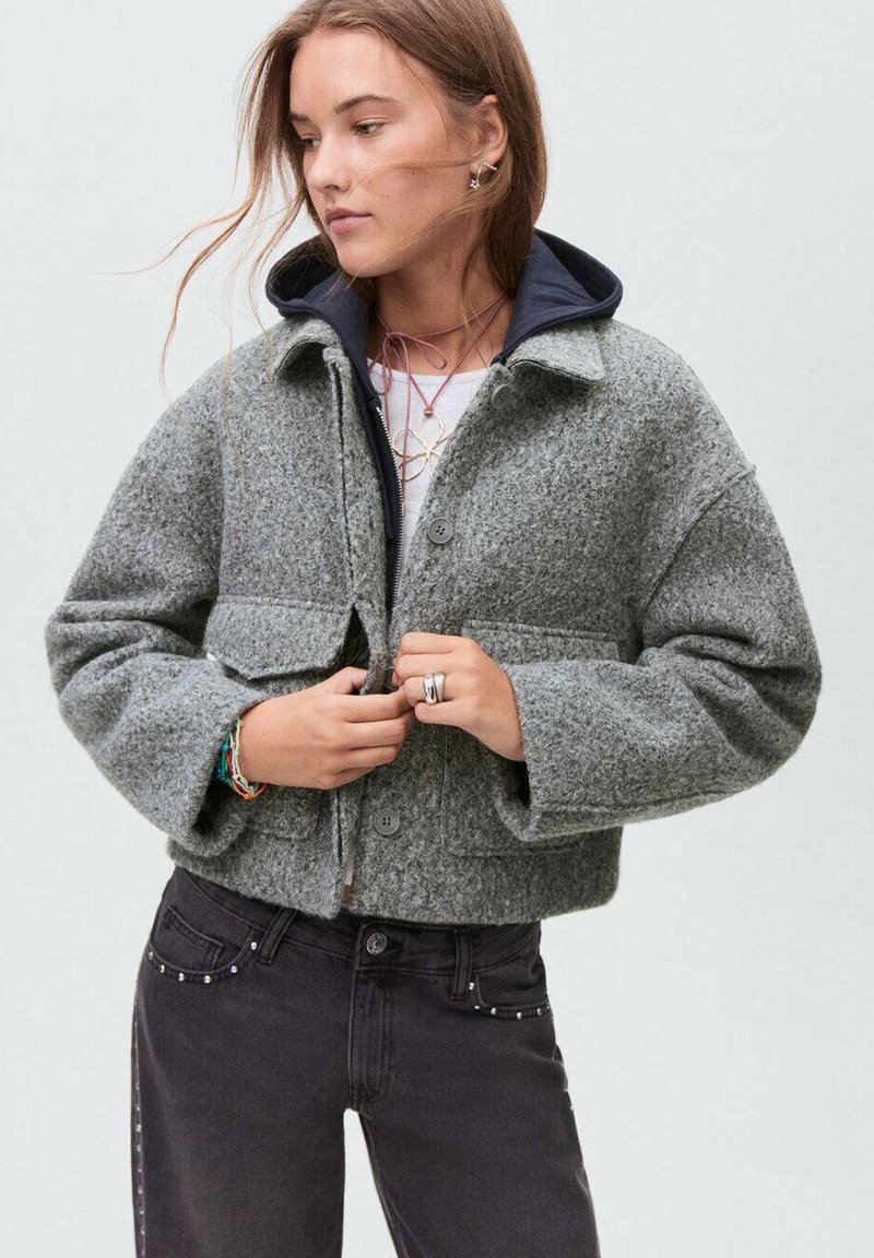 Veste en laine gris coupée avec fermeture à bouton, dotée de deux poches avant et d'une capuche bleu marine. Associée avec un jean noir orné de clous.