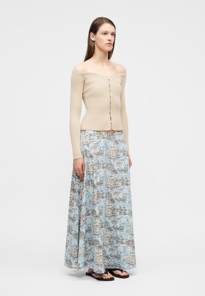 Γυναίκα που φοράει μπεζ ριμπ μπλούζα ώμου με κουμπιά εμπρός, μπλε φούστα μάξι με σχέδιο και μαύρα σαγιονάρες, στέκεται μπροστά σε λευκό φόντο.