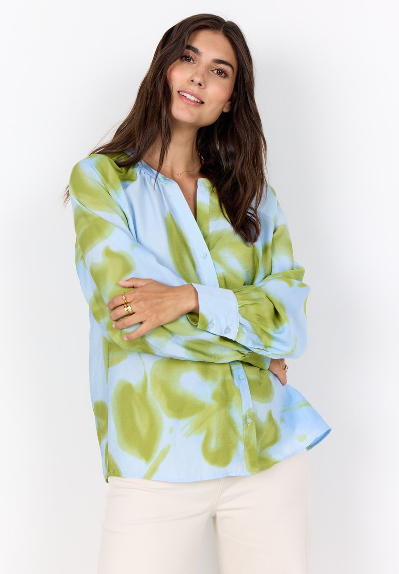 Femme souriante aux longs cheveux bruns portant une chemise ample bleu et vert tie-dye à boutons et un pantalon clair, bras croisés.