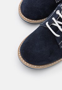 Marineblaue Wildlederschuhe mit weißen Schnürsenkeln, ausgestattet mit einer gesteppten, hellbraunen Sohle und einem runden Zehendesign. Strukturiertes Obermaterial für besseren Halt.