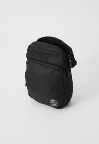 Mochila negra de nailon con forma rectangular, cierre de cremallera, correa ajustable y un parche de marca en la parte frontal. Superficie texturizada.