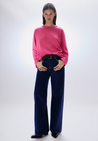 Rózsaszín-fehér csíkos hosszú ujjú ing, sötétkék, magas derekú, széles szárú farmernadrággal, amelyen ezüst szegecsek díszítik az oldalakat.