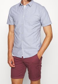 Košile s krátkým rukávem v světle modré barvě s jemným tečkovaným vzorem, spárovaná s burgundskými šortkami, které mají ohrnuté lemy a boční kapsy.