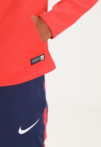 Punainen urheilu-paita pitkähihaisena, jossa on tasku ja logo-patch; yhdistettynä tummansinihin shortseihin, joissa on kuvio ja valkoinen brändäysyksityiskohta.