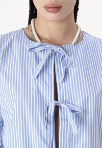 Blouse rayée bleue avec un col rond, dotée de deux liens en ruban et d'une légère ouverture à l'avant, accompagnée d'un collier de perles.