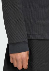 Dark gray long-sleeve top featuring a textured waffle knit design. Cuffs are solid and fitted, blending with the overall fabric structure.