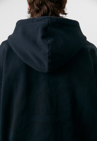 Felpa nera con cappuccio caratterizzata da un tessuto testurizzato con un design grafico prominente sulla schiena, cuciture ben definite e un cappuccio strutturato.