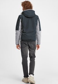 Herrejakke i mørk marineblå og lys grå, har hette, elastisk midje og justerbare mansjetter. Kombinert med grå jeans og sneakers.