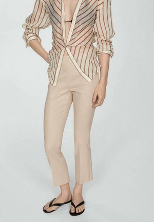 Personne portant un pantalon beige raccourci, une chemise rayée à boutons partiellement ouverte et des tongs noires, debout avec une main sur la hanche.