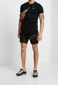 Man wearing black t-shirt, camo shorts, black-and-white sneakers, and a brown crossbody bag, standing against a plain light background.