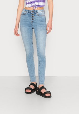 Person wearing light blue high-waisted skinny jeans with button fly, purple tie-dye crop top, and black open-toe sandals.