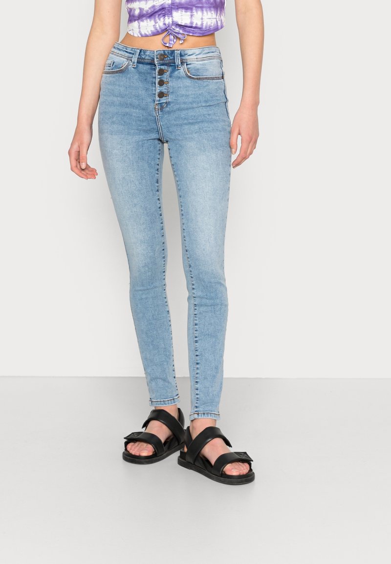 Osoba ubrana w jasnoniebieskie, obcisłe jeansy z wysokim stanem i zapięciem na guziki, fioletowy top crop o wzorze tie-dye oraz czarne sandały z odkrytymi palcami.