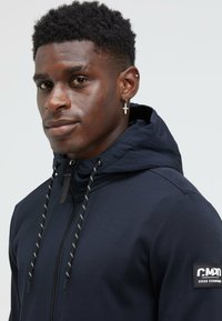 Navy hoodie with a zip front, adjustable drawstrings, and a branded patch on the sleeve. Smooth fabric with a fitted design.