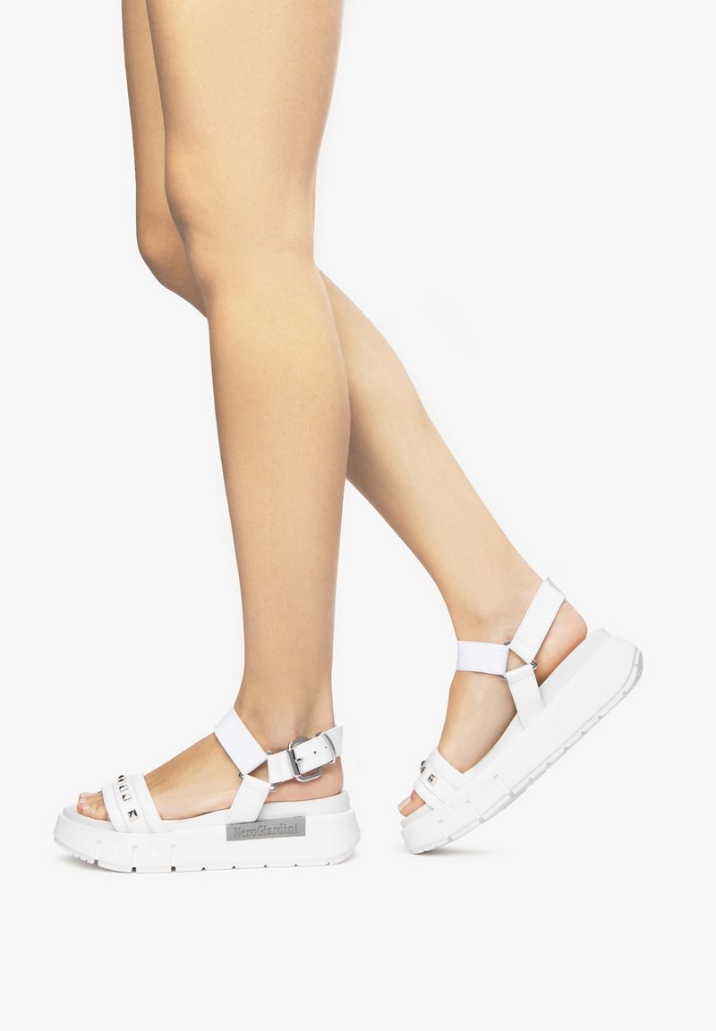 Sandales à plateforme blanches avec semelle texturée et bride de cheville ajustable. Des accents de boucle rehaussent le design. Portées sur des jambes bronzées sur un fond blanc.
