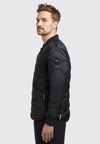 Black puffer jacket with a textured, quilted design featuring rounded shapes. Includes a black collar and side pockets with subtle branding.