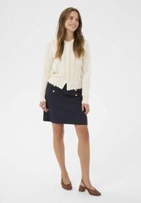 Gaišs krēma krāsas adīts džemperis ar ribotu tekstūru, viļņotu apakšmalu un pogu aizdari. Tumši zils pildspalvas svārki ar zelta pogu akcentiem. Brūni austie slingback papēži.