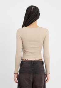 Ilga rankovėmis smėlio spalvos ribota palaidinė su sutrumpintu dizainu ir banguotu kraštu, derinama su tamsiai mėlynomis kelnėmis, turinčiomis dideles galines kišenes.