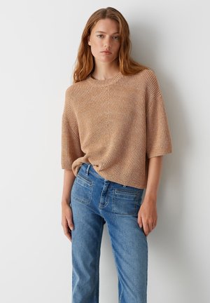 Helles beige Strickoberteil mit kurzen Ärmeln, strukturierter Musterung und rundem Halsausschnitt, kombiniert mit dunkelblauen Jeans in mittlerer Leibhöhe mit Gesäßtaschen.