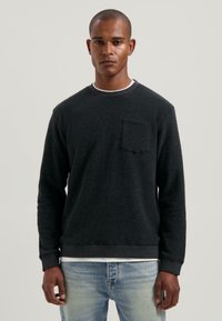 Giovane uomo in piedi, che indossa un maglione nero a maniche lunghe con taschino su una camicia bianca e jeans azzurri chiari, su uno sfondo neutro.