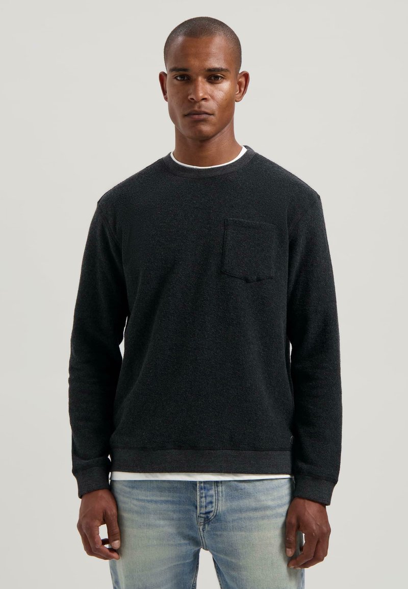 Giovane uomo in piedi, che indossa un maglione nero a maniche lunghe con taschino su una camicia bianca e jeans azzurri chiari, su uno sfondo neutro.