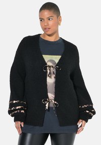 Černý pletený cardigan s výstřihem do "V", leopardími potisky na rukávech a předními šňůrkami. Texturovaná látka dodává hloubku.