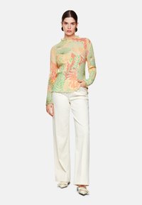 Leichtes, langärmeliges Oberteil aus transparenter Stoff mit einem floralen Muster in Grün und Orange. Kombiniert mit weißen, weit geschnittenen Hosen und flachen Schuhen.