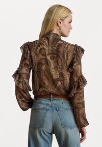 Brun paisley-mønstret bluse med volangærmer, med høy hals, kombinert med lyseblå høytaljede jeans og et belte.