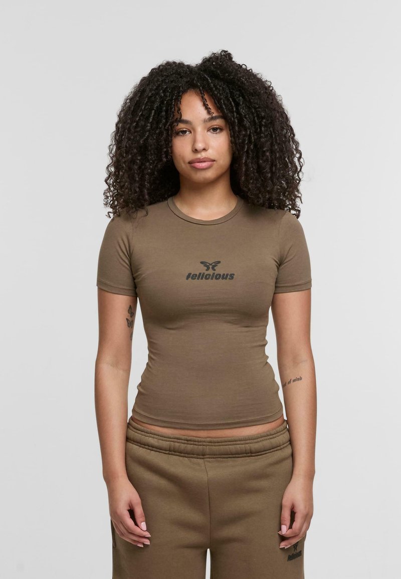 Braunes, tailliertes T-Shirt mit kurzen Ärmeln und einem schwarzen "felicious"-Logo auf der Brust. Getragen mit passenden hellbraunen Jogginghosen.