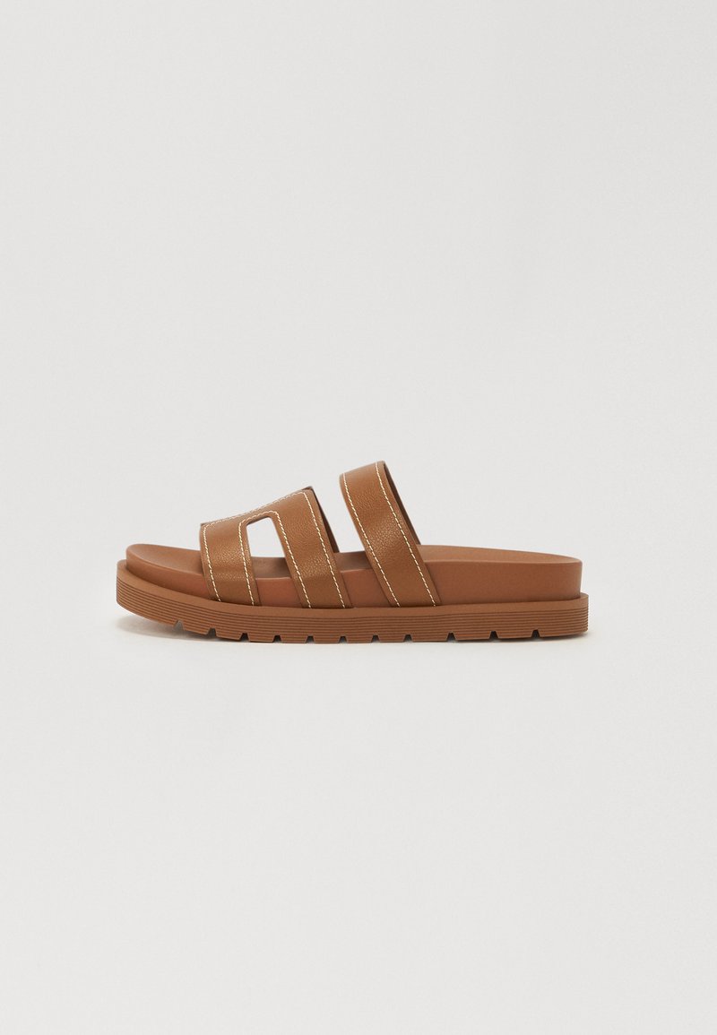 Brune skyvesandaler med to brede stropper, teksturert materiale og en tykk gummisåle. Har kontrasterende hvit søm og flatt design.