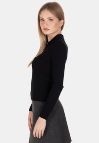 Giovane donna con lunghi capelli biondi che indossa un top nero a coste con maniche lunghe e una gonna grigia, in posa di lato su uno sfondo bianco.