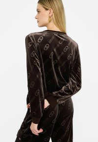Hnedý zamatový loungewear set s jemným vzorom loga. Obsahuje dlhé rukávy, rebrované manžety a uvoľnený strih. Mäkkučká textúra.