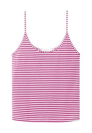 Femme debout, les mains dans le dos, portant un débardeur rayé rose et blanc, un pantalon large bleu marine et des sandales roses avec une boucle devant un mur blanc.