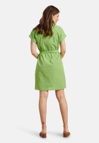 Kurzes grünes Kleid mit aufgerollten Ärmeln und einem Bindegürtel in der Taille. Der Stoff wirkt leicht und hat eine glatte Textur.