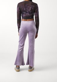 Person som bär lavendelvelour byxor med vida ben, svart och lila långärmad crop-top, och grå sneakers, som står framför en vit bakgrund.