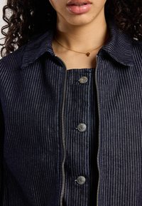 Marine gestreepte jas met een voorrits en drukknoopsluitingen. Bevat een kraag en een gladde textuurstof. Gouden ketting zichtbaar.
