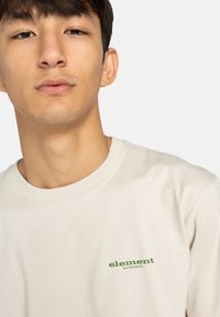 Helle beige Baumwoll-T-Shirt mit Rundhalsausschnitt und kurzen Ärmeln. Mit grünem "element" Logo auf der linken Brustseite. Glatte Textur.