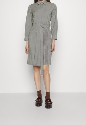 Vrouw draagt een knielange, lange mouwen, geruite jurk met een taille riem, bruine sokken en donkerbruine loafers met gouden gespen.