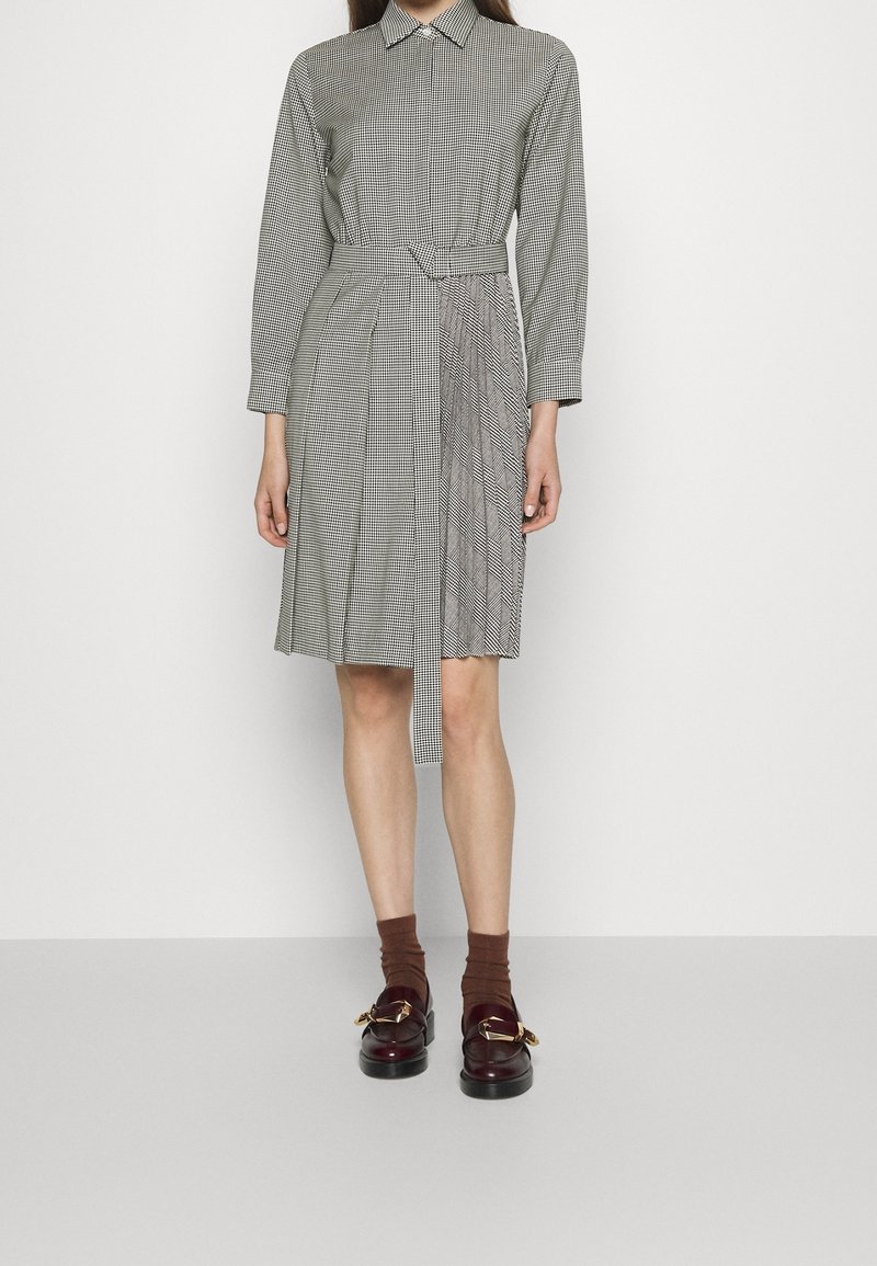 Femme portant une robe à carreaux mi-longue, manches longues, avec une ceinture à la taille, des chaussettes marron et des mocassins marron foncé avec des boucles dorées.
