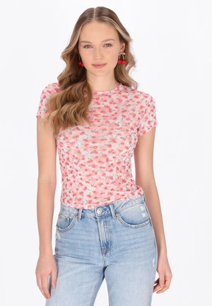 Młoda kobieta o falowanych blond włosach, ubrana w różową bluzkę z krótkim rękawem w wzór z wisienkami, jasnoniebieskie dżinsy z wysokim stanem oraz kolczyki w kształcie wisienek.