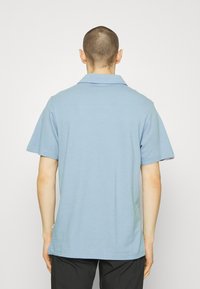 Osoba s krátkými vlasy, oblečená v světle modrém polo tričku s krátkým rukávem a černých kalhotách, otočená zády k prostému bílému pozadí.