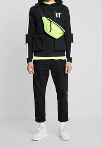 Osoba v černé mikině s bílými pruhy, černých kalhotách, bílých teniskách a neonově zelené tašce přes rameno a vrstvě trička.