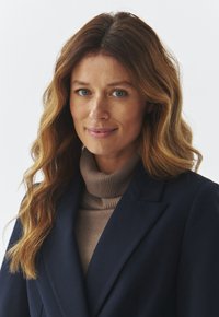 Žena s dugom, valovitom smeškom kestenjaste kose, obučena u usku tamnoplavu jaknu preko rebrastog taupe turtleneck-a, na beloj pozadini.