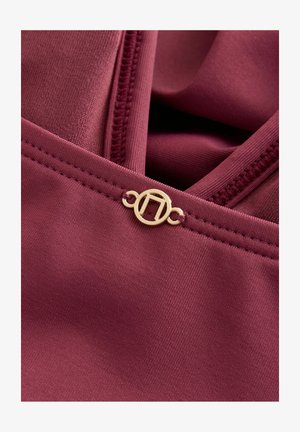 Maronowy materiał o gładkiej teksturze z złotym okrągłym akcentem w pobliżu zamka błyskawicznego. Projekt zawiera subtelne detale szycia.